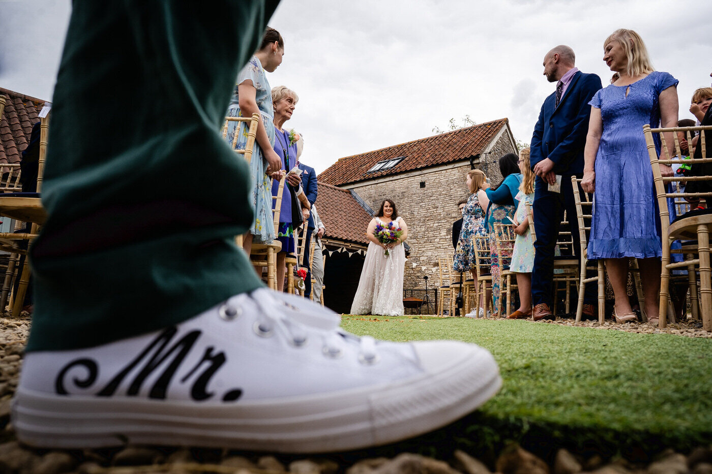 Guests stand for an outdoor barn ceremony while the bride waits at the end of the aisle, framed past a groom's white trainer marked Mr.