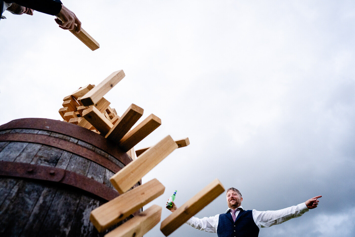A giant Jenga tower topples off a barrel against a grey sky while a laughing guest in a waistcoat throws both arms wide.